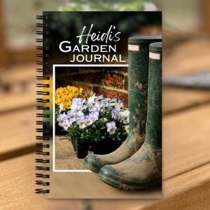 May include: A spiral-bound journal titled "Heidi's Garden Journal." The cover features a photograph of green rain boots, potted flowers, and the journal's title in white and black text. The journal is on a wooden surface.