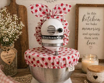 May include: A white stand mixer featuring a pink fabric cover with a red strawberry pattern. The cover includes a ruffled base for the bowl and a top section with two upright, bunny-ear-shaped fabric pieces. The mixer has a silver metal bowl.