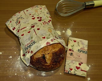 May include: A reusable food wrap bag featuring a cream background with a pattern of red cherries, hearts, whisks, and chef hats, printed with the text Bless the BAKER. A folded matching wrap is beside a round loaf of bread and a metal whisk.