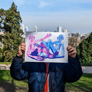 Peut inclure: Une personne tient un dessin de deux personnages, un rose et un bleu, avec des membres allongés. Les personnages sont entrelacés sur un fond blanc. Le dessin est tenu devant la personne, avec des arbres et des bâtiments en arrière-plan.