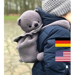 May include: A gray crocheted teddy bear backpack worn by a child wearing a blue jacket and a gray knitted hat. The child is standing in front of a green background.