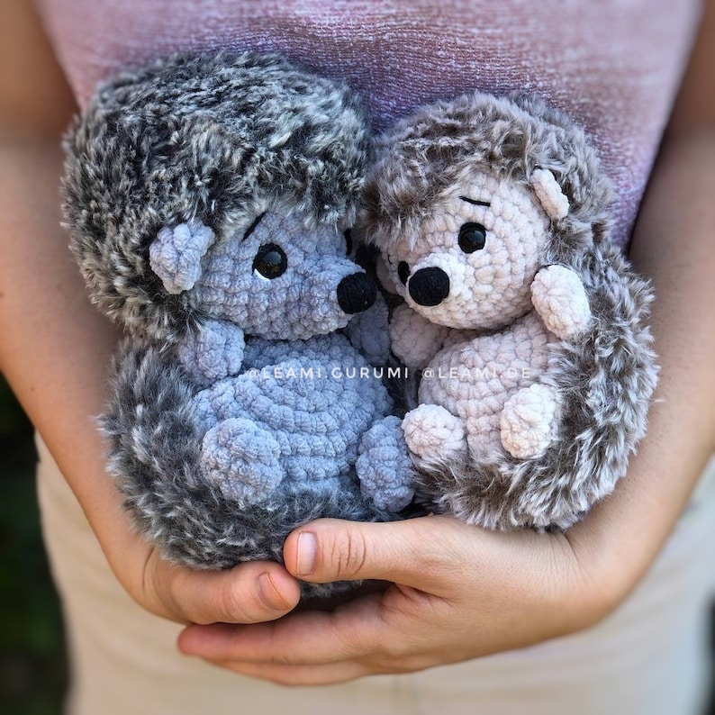 May include: Two crocheted hedgehog plushies, one gray and one beige, are held in a person's hands. The hedgehogs have black eyes and noses and are made with a soft, fuzzy yarn.
