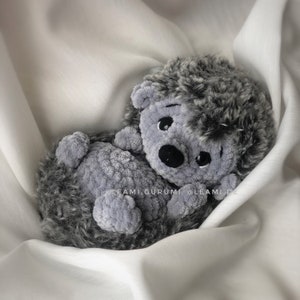 May include: A gray and black crocheted hedgehog plush toy with a sweet expression. The toy is lying on a white fabric background.