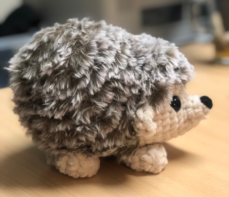 May include: A brown and white crocheted hedgehog plush toy with black eyes. The hedgehog is sitting on a wooden surface.