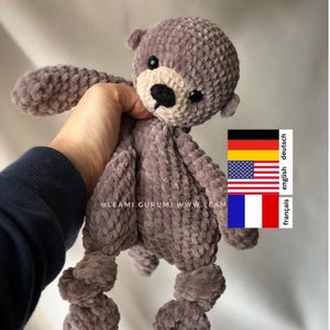 Può includere: Un giocattolo di peluche all'uncinetto a forma di lontra. La lontra è grigia con il naso marrone e gli occhi neri. Il giocattolo è tenuto da una mano.