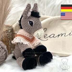 Puede incluir: Un juguete de peluche de burro gris tejido a crochet con pezuñas negras y una bufanda tejida a crochet rosa y naranja. El burro está sentado sobre una superficie blanca y esponjosa. El texto "www.leami.de @leami_crumi" es visible en el juguete.