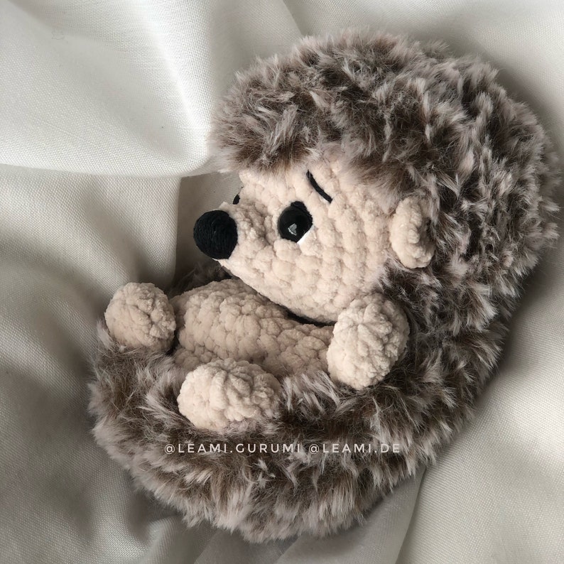 May include: A crocheted hedgehog plush toy with brown fur and a beige body. The hedgehog is curled up in a ball with its eyes closed. The toy is sitting on a white fabric background.