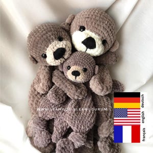 Op de afbeelding: Drie gehaakte pluche zeehondjespeelgoed in een knuffel. De zeehonden zijn bruin en hebben zwarte ogen en neuzen. De grootste zeehond is achteraan, de twee kleinere zeehonden zijn ervoor. De zeehonden knuffelen elkaar allemaal.