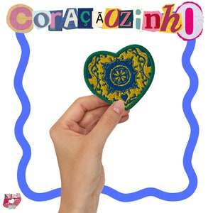 Könnte beinhalten: Eine Hand hält ein herzförmiges Patch in Grün und Gelb mit einem blauen und gelben Blumenmuster in der Mitte. Das Wort "Corazón" ist in bunten Buchstaben über dem Herzen geschrieben.