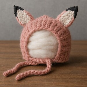 Può includere: Un cappello per neonati lavorato a maglia con un design a volpe. Il cappello è principalmente rosa polveroso con orecchie interne color crema e punte nere. Il cappello ha due lunghe cravatte ed è progettato per un neonato.