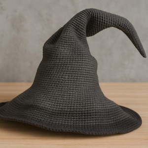 May include: A dark grey witch's hat with a wide brim and a pointed, curved tip. The hat is made of a textured fabric, possibly knitted or crocheted, and is displayed on a wooden surface. A Halloween costume accessory.