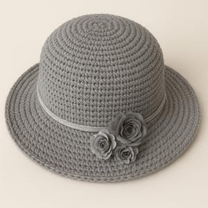 Op de afbeelding: Een grijze gehaakte hoed met een brede rand en een decoratieve band. Drie grijze stoffen rozen sieren de zijkant en voegen elegantie toe. De hoed is een stijlvol accessoire.