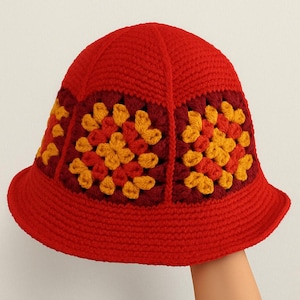 Può includere: Un cappello a secchiello rosso all'uncinetto con una tesa larga. Il cappello presenta pannelli quadrati con un motivo floreale nei toni del giallo e del bordeaux. Il cappello è fatto a mano e ha un aspetto strutturato.