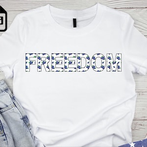 May include: White t-shirt with the word "FREEDOM" printed across the front. The letters are outlined in black and filled with a blue floral pattern. The shirt is paired with blue jeans and an American flag.