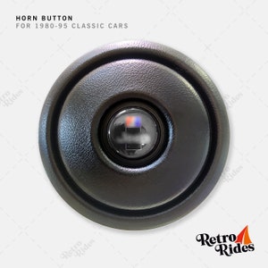 May include: Black round horn button for classic cars, 1980-95. The button has a black plastic cover with a circular opening in the center. The opening reveals a red and blue button.