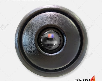 May include: Black round horn button for classic cars, 1980-95. The button has a black plastic cover with a circular opening in the center. The opening reveals a red and blue button.