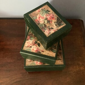 Puede incluir: Tres cajas de madera cuadradas apiladas, con marcos verde oscuro y tapas adornadas con diseños florales de rosas. Las rosas son rosas y blancas con follaje verde, sobre un fondo beige.