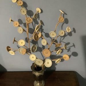 May include: A decorative twig arrangement with various sized buttons in shades of beige, brown, and blue, displayed in a silver vase.