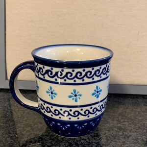 Könnte beinhalten: Ein Keramikbecher mit cremefarbener Basis, dunkelblauem Rand und Henkel. Die Außenseite ist mit horizontalen Bändern aus dunkelblauen Wellenmustern, Reihen kleiner Punkte und einer mittleren Reihe hellblauer Blumenmotive verziert.