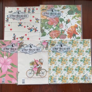 May include: Five packages of folded gift wrap sheets in clear plastic sleeves. Each package features the text GIFTWRAP/EMBALLAGE CADEAU and contains two sheets. Designs include clowns with hearts, orange roses, pink lilies, a couple on a bicycle, and yellow flowers.