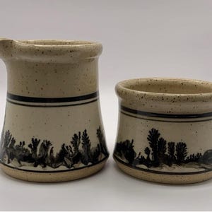 Puede incluir: Un juego de jarra de crema y azucarero de cerámica de color crema con un diseño de escena de bosque en blanco y negro.
