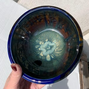 May include: A handcrafted ceramic bowl with a glossy finish. The bowl has a deep blue rim and a centre with shades of teal, brown, and cream. The interior has a unique, swirling pattern, creating an artistic and functional piece.