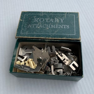 Puede incluir: Una caja verde vintage abierta, etiquetada como "ROTARY ATTACHMENTS", contiene varios accesorios plateados y crema para máquinas de coser. La caja rectangular con tapa con bisagras está llena de prensatelas de metal y otros accesorios de costura.