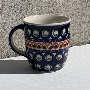 May include: A ceramic mug featuring a dark blue base colour with a repeating pattern of white and green circles. A central horizontal band displays a row of orange and black leaf-like shapes. The interior of the mug is a solid off-white colour.