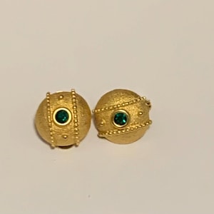 Könnte beinhalten: Ein Paar runde, goldfarbene Ohrringe mit einer texturierten, matten Oberfläche. Jeder Ohrring verfügt über einen zentralen, kreisförmigen smaragdgrünen Edelstein in einer Goldfassung, flankiert von zwei vertikalen Perlenlinien und kleinen Punkten.