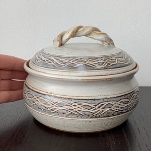 以下が含まれることがあります： クリーム色の釉薬とブルーグレーのアクセントが施された、蓋付きの丸い陶器製ジャー。蓋には縄状のツイストハンドルが付いており、蓋と本体の両方に複雑な編み込み模様のエンボス加工が施されています。