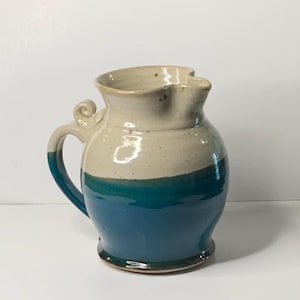 May include: A handmade ceramic pitcher with a cream-colored top and a teal blue base. The pitcher has a handle and a decorative swirl detail. The base is a darker teal blue, and the rim has a unique, wavy shape.