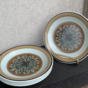 Puede incluir: Un juego de cuatro platos de cerámica con un diseño floral. Cada plato presenta un centro marrón con un patrón floral turquesa, rodeado por un borde azul claro y marrón. Los platos están apilados y expuestos.