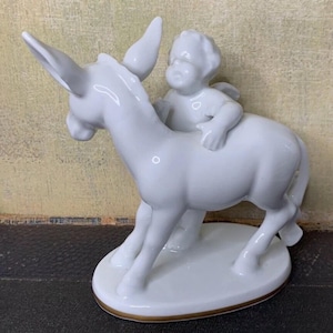 Puede incluir: Una figura de porcelana blanca que representa a un querubín montado en un burro. El querubín tiene pequeñas alas y está sentado en la espalda del burro. El burro tiene orejas grandes y está sobre una base redonda con un borde dorado.