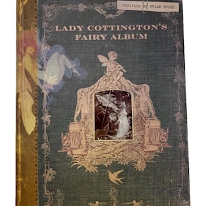 May include: A vintage book titled "Lady Cottington's Fairy Album" with a teal cover and gold accents. The cover features illustrations of fairies, a central framed image, and the text "Verified by Brian Froud".