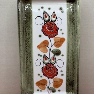 Op de afbeelding: Rechthoekige keramische serveerschaal met een groen gespikkelde rand en een witte midden. Het midden heeft een bloemenontwerp met rode rozen, oranje bladeren en groene accenten. De schaal heeft aan elk uiteinde een handvat.