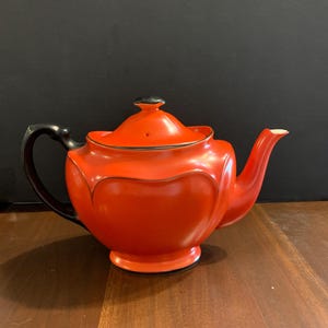 May include: A bright orange teapot featuring a smooth, rounded body with scalloped edges, a black handle, a black lid knob, and a black base. The spout is orange with a white interior tip, and the lid has a small steam hole near the centre.
