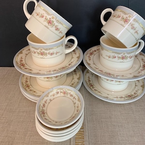 Peut inclure: Un ensemble de tasses à thé et soucoupes en céramique crème, ornées d'un motif floral délicat de roses roses et de feuilles vertes, rehaussé d'une bande bleu doux et dorée sur les bords. La collection comprend des tasses et soucoupes empilées.