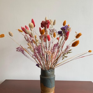 May include: A bouquet of dried flowers in shades of red, orange, purple, and cream arranged in a cylindrical ceramic vase. The vase features a two-tone design with a dark blue-grey upper section and a textured, earthy brown base.