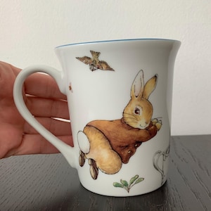 Puede incluir: Una taza de cerámica blanca con una ilustración colorida de un conejo marrón con chaqueta, representado en movimiento de salto. Sobre el conejo, un pequeño pájaro vuela y un brote de planta verde es visible cerca de la base de la taza.