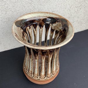 Puede incluir: Un jarrón de cerámica hecho a mano con un diseño único. El jarrón presenta un borde ancho y acampanado y un cuerpo cilíndrico con cortes verticales. La combinación de colores incluye tonos de marrón, beige y un toque de esmalte marrón oscuro brillante.