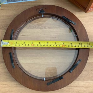 May include: A circular brown wooden picture frame with a glass center, featuring black metal swivel clips on the back. A yellow measuring tape is placed across the frame, showing a diameter of approximately 11.5 inches from edge to edge.