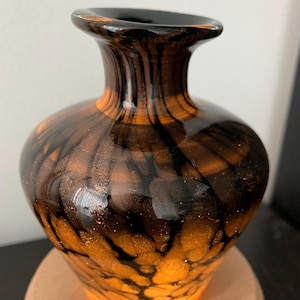May include: A small glass vase with a bulbous body and a flared rim. The glass features a swirling pattern of deep black and translucent amber tones, accented with fine gold-coloured glitter particles embedded throughout the material.