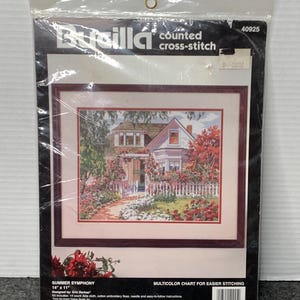 Puede incluir: Un kit de punto de cruz contado Bucilla sellado, titulado "Summer Symphony". La imagen muestra un punto de cruz enmarcado de una casa con una valla blanca y flores coloridas. El kit mide 35,56 cm x 27,94 cm.