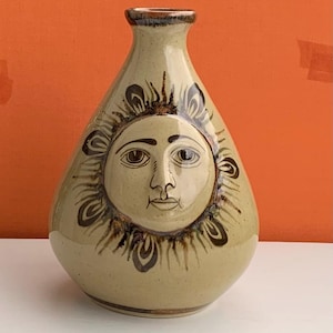May include: A ceramic vase featuring a sun face design. The vase is a light beige colour with a brown rim and sun details. The sun face has brown eyes, a nose, and a mouth. The vase is teardrop-shaped.