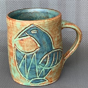Pode incluir: Uma caneca de cerâmica com um design de pássaro azul esculpido à mão sobre uma superfície texturizada com esmalte castanho e verde manchado. A caneca tem uma pega robusta e uma forma cilíndrica, com um acabamento rústico e textura visível.