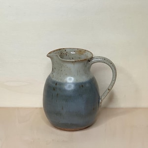 Può includere: Una brocca in ceramica fatta a mano con un corpo arrotondato e un manico. La brocca è bicolore, con una parte superiore grigio chiaro e una parte inferiore blu-grigio più scura. Ha un beccuccio per versare ed è adatta per servire bevande.