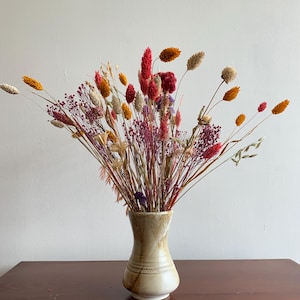 Puede incluir: Un jarrón de cerámica con esmalte moteado en beige y marrón que contiene un vibrante arreglo de flores y hierbas secas en tonos rosa, naranja, amarillo y crema. El jarrón presenta un cuello estrecho y una banda texturizada en la base.