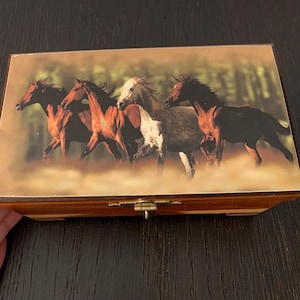 May include: A rectangular wooden box featuring a printed image on the lid of four horses running across a field. The horses are depicted in shades of brown, black, and white. The box has a gold-coloured metal latch centred on the front edge.