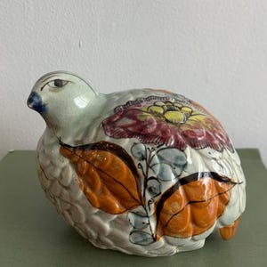 May include: A ceramic bird figurine with a crackled off-white glaze, featuring a large painted red and yellow flower on its back, orange leaf motifs along the side, and a dark blue beak, showcasing a textured surface with visible fine lines.