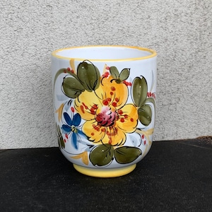 May include: A white ceramic vase with a yellow base and rim, featuring a large yellow flower with red and orange details, green leaves, and small blue flowers. The hand-painted floral design adds a touch of artistry to this decorative piece.